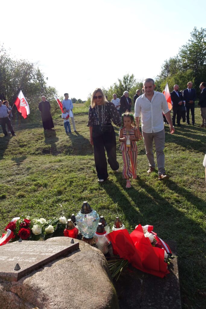 Grupa osób na trawie z flagami Polski, kwiaty i znicze na kamieniu z tablicą pamiątkową, drzewa w tle, słońce oświetla scenę od tyłu.