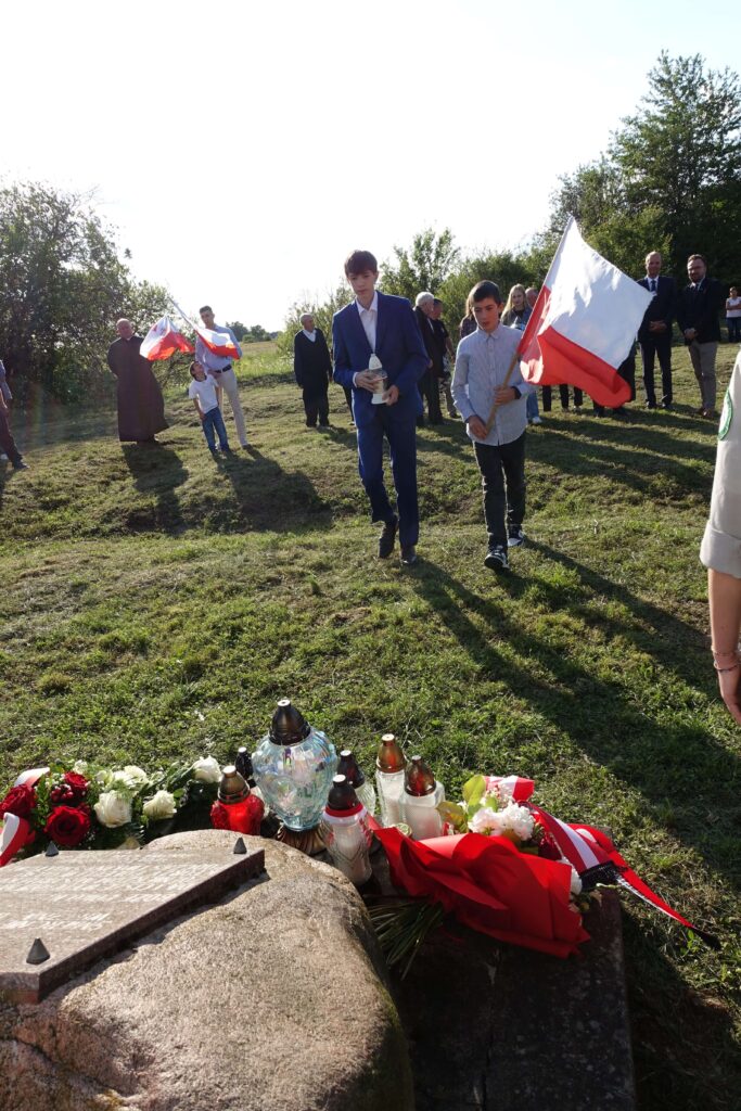 Kamień nagrobny z tablicą, znicze i kwiaty z biało-czerwonymi wstęgami, w tle grupa ludzi, dwóch chłopców z flagą Polski, trawiasty teren, drzewa.