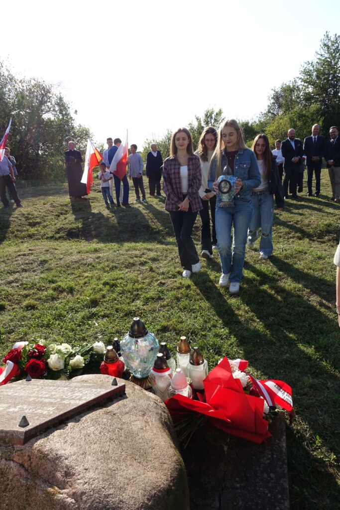 Grupa ludzi na łące, czworo młodych kobiet idzie z przodu, jedna niesie lampion, w tle flagi, kwiaty i znicze na kamieniach.