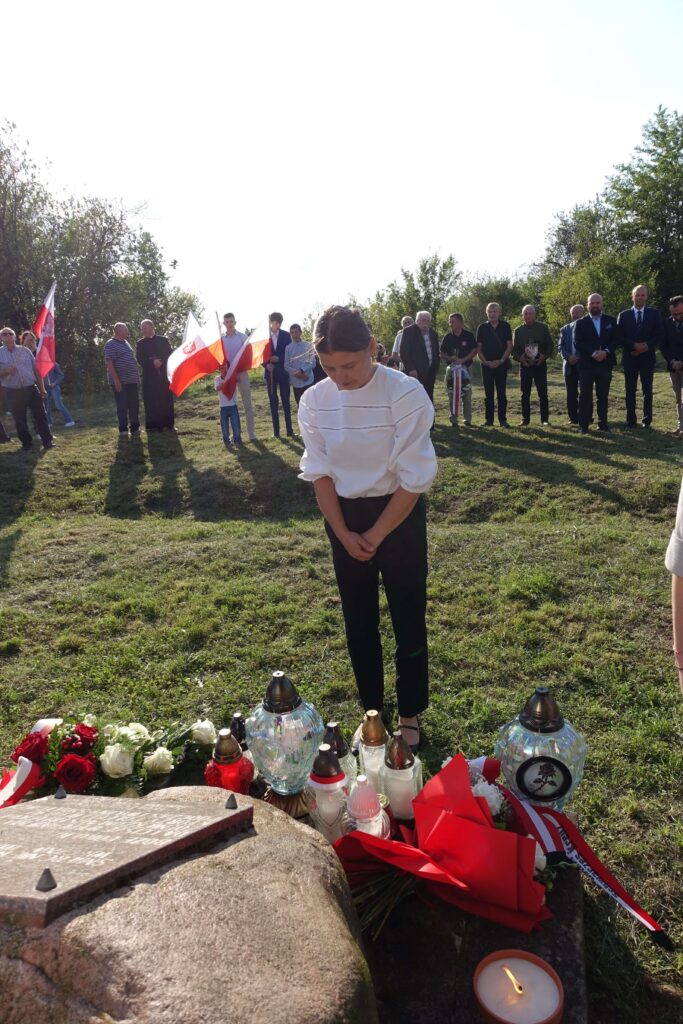 Kobieta w białej bluzce i czarnych spodniach stoi na trawie przed kamieniem z tablicą, wokół znicze, kwiaty i wieńce, w tle ludzie z biało-czerwonymi flagami.