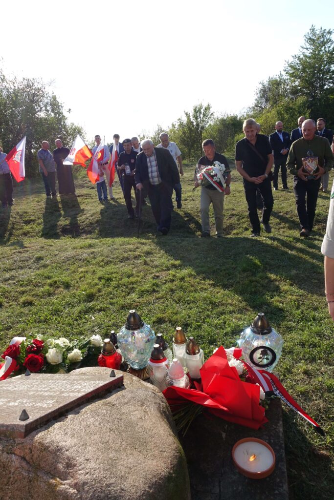 Grupa ludzi idących po trawie ku kamieniowi z tablicą, na pierwszym planie znicze, czerwono-białe kwiaty i zapalona świeca, w tle drzewa i kilka osób z flagami Polski.