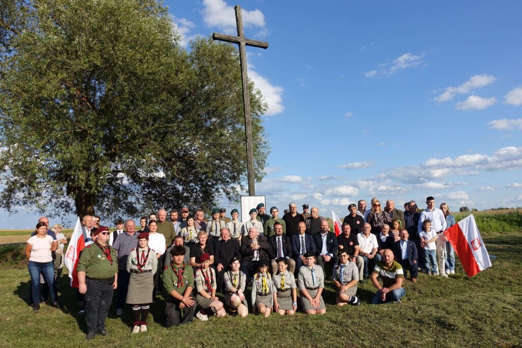 Grupa ludzi w różnym wieku, w tym harcerze w mundurach, stoją i siedzą na trawie przed dużym drewnianym krzyżem i drzewem, pod niebieskim niebem z chmurami.