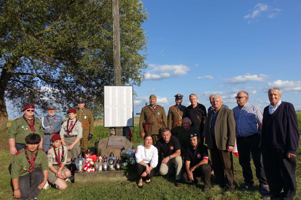 Grupa ludzi w mundurach harcerskich i wojskowych, kilku w cywilnych ubraniach, stojących i klęczących przy pomniku z tablicą i zniczami, na tle zieleni i nieba.