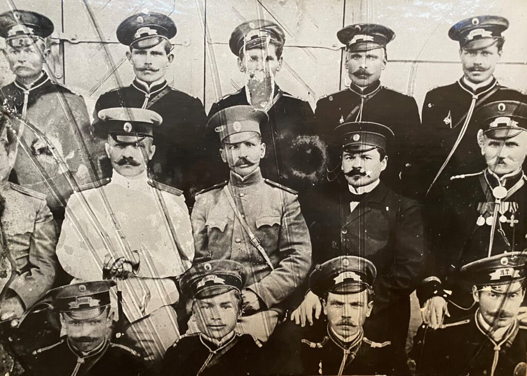 Czarno-biała fotografia grupowa mężczyzn w mundurach wojskowych i czapkach z daszkiem, niektórzy z wąsami, widoczne zarysowania i uszkodzenia zdjęcia.