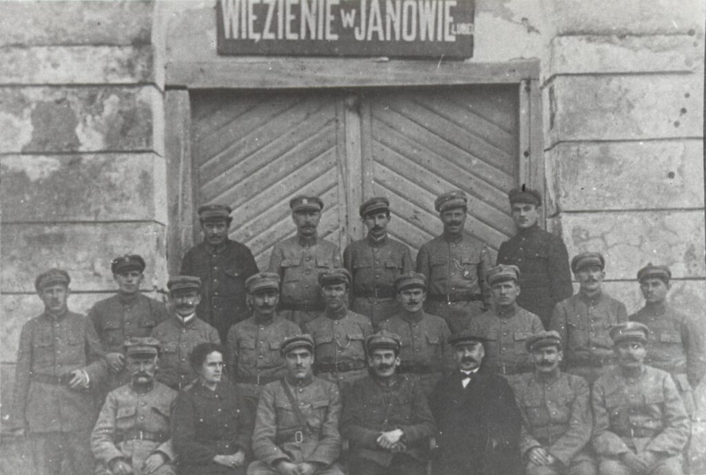 Grupa mężczyzn w mundurach i czapkach przed drewnianymi drzwiami budynku z napisem "Więzienie w Janowie".