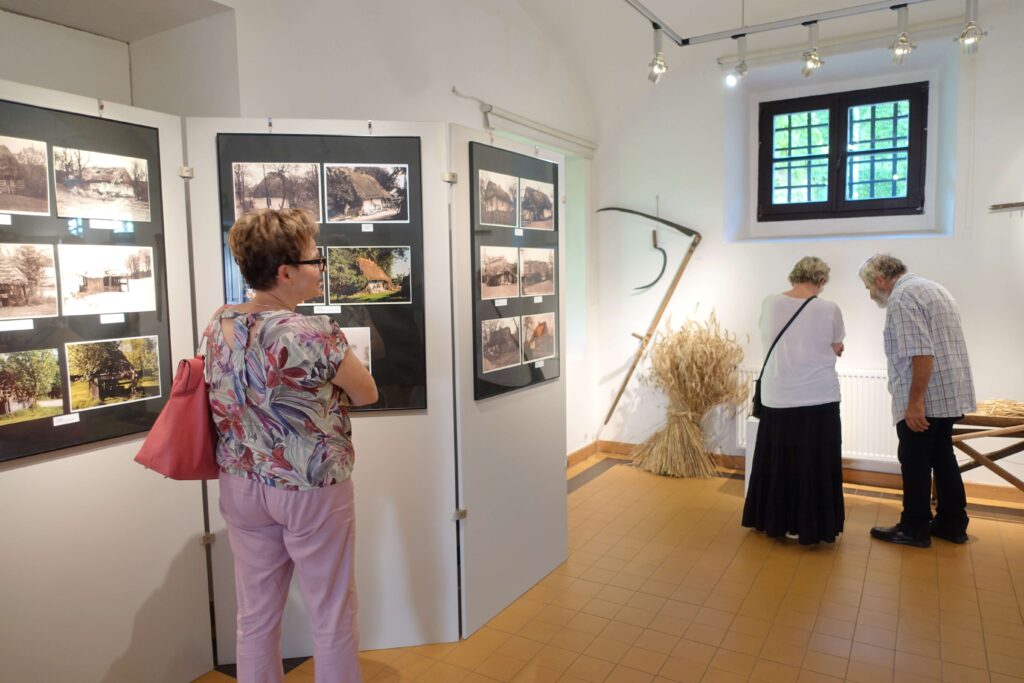 Trzy osoby w jasnym pomieszczeniu, dwie oglądają eksponaty na ścianach z fotografiami chat, jedna para pochylona nad czymś na stole, na ścianie kosa i snop zboża.