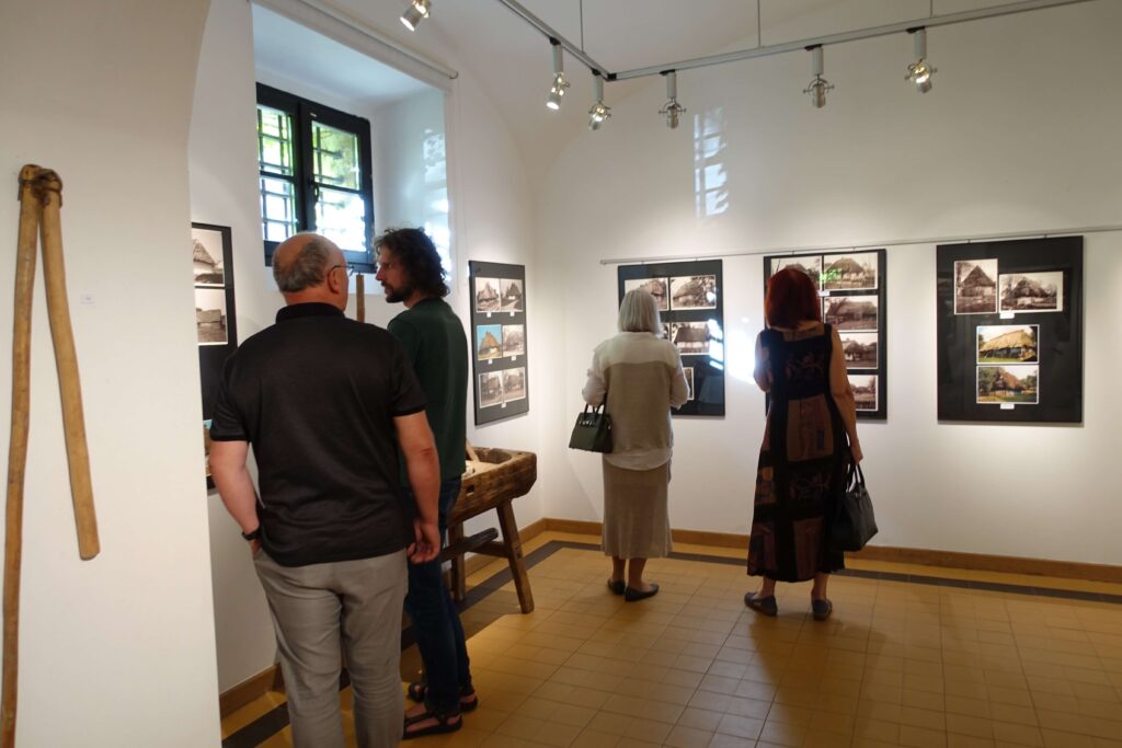 Wnętrze galerii z białymi ścianami, czterema osobami oglądającymi czarno-białe fotografie na ścianach, żółto-brązowa podłoga, okno z kratami, drewniany przedmiot.