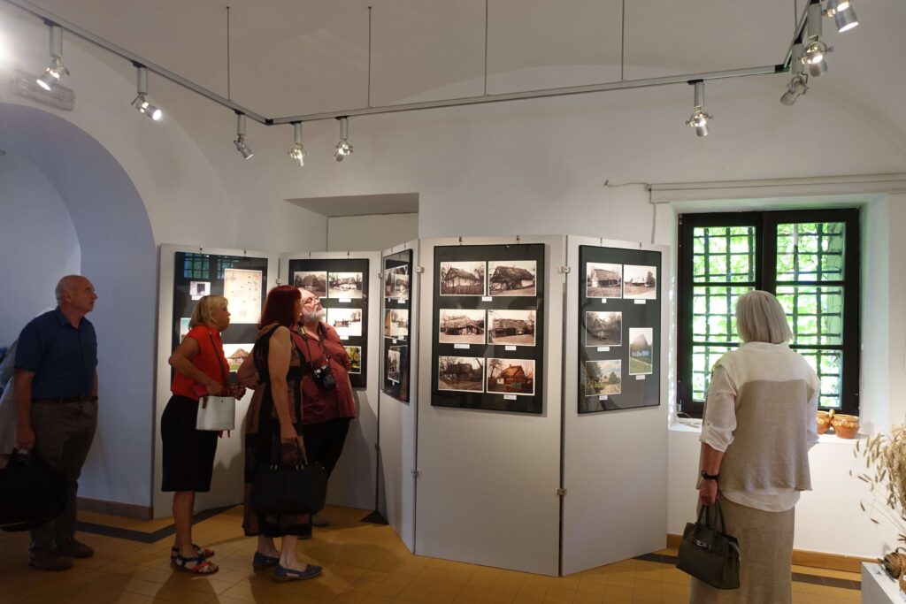Grupa ludzi stoi w jasnym wnętrzu galerii z białymi ścianami, oglądając plansze z czarno-białymi fotografiami budynków, obok okno z kratą i zielonymi liśćmi za szybą.