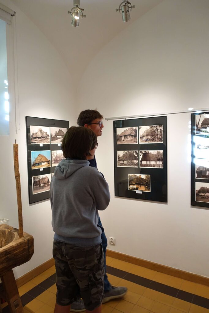 Dwóch chłopców stoi przed ścianą z czarnymi ramami zawierającymi fotografie starych, wiejskich domów, jasne ściany, żółto-brązowa podłoga.