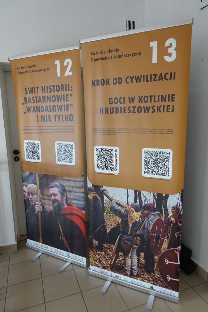 Dwa stacjonarne roll-upy z brązowym tłem, tekstem w języku polskim i zdjęciami ludzi w historycznych strojach trzymających tarcze i broń, stojące na jasnej podłodze przy białej ścianie.