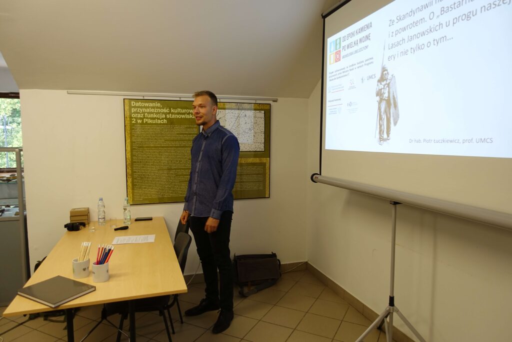 Mężczyzna w granatowej koszuli stoi przy stole z butelkami, kubkami z ołówkami i dokumentami, obok ściany z planszą i rzutnika wyświetlającego tekst i grafikę.