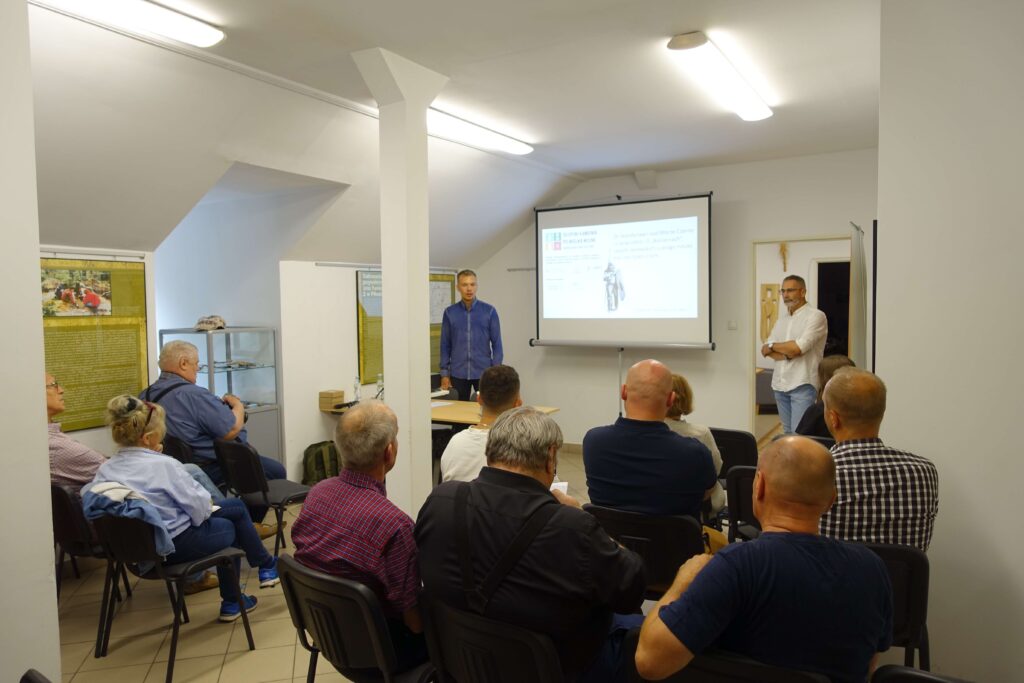 Sala z białymi ścianami i sufitami, ludzie siedzący na czarnych krzesłach, mężczyzna w niebieskiej koszuli stoi przy stoliku, biały ekran z projekcją, oświetlenie sufitowe.