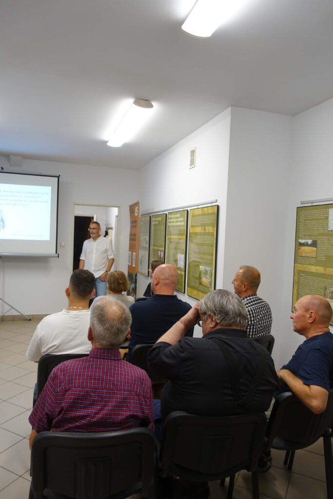 Mężczyzna w białej koszuli stoi obok ekranu projekcyjnego, przed grupą siedzących osób, w jasnym pomieszczeniu z białymi ścianami ozdobionymi tablicami informacyjnymi.