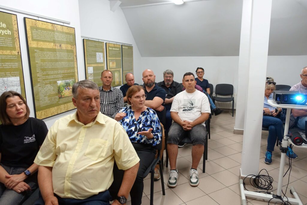 Grupa osób siedzących na krzesłach w jasnym pomieszczeniu z tablicami informacyjnymi na ścianie, projektor na statywie po prawej stronie.