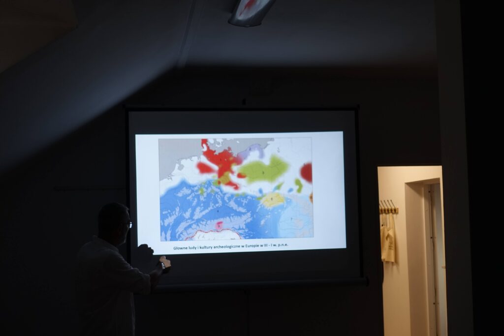 Ekran z mapą Europy z kolorowymi zaznaczeniami, zarys sylwetki mężczyzny, ciemne pomieszczenie, jasny korytarz z wieszakami i torbami.