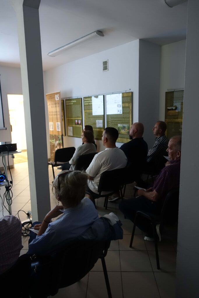 Grupa osób siedzących na krzesłach w ciemnym pomieszczeniu, na ścianie plansze informacyjne i baner z tekstem, podłoga jasne płytki.