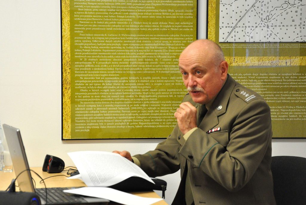 Starszy mężczyzna w mundurze wojskowym siedzi przy stole z dokumentami i laptopem, w tle zielona plansza z tekstem i mapą.
