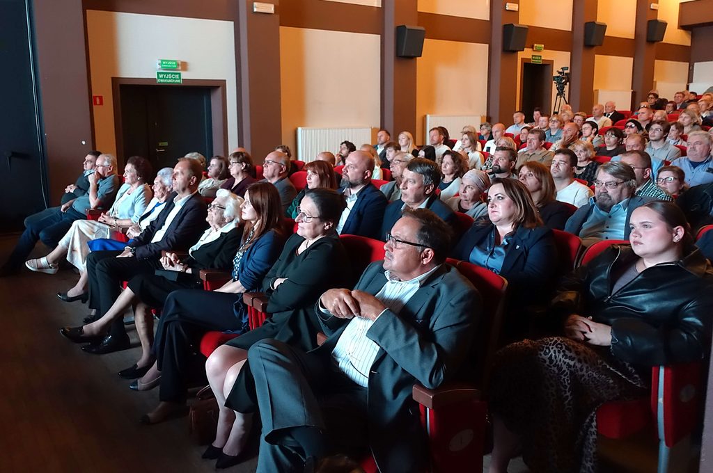 Wnętrze sali z czerwonymi fotelami, wypełnione siedzącymi osobami w różnym wieku, ściany brązowo-beżowe, kamera na statywie z tyłu.