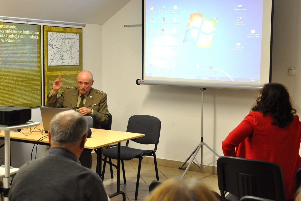 Mężczyzna w mundurze wojskowym siedzi przy stole z laptopem, obok ekran projekcyjny z widocznym pulpitem Windows, na pierwszym planie osoby siedzące tyłem.