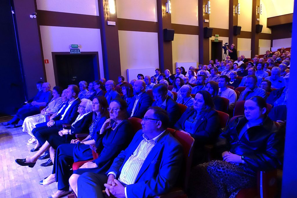 Ludzie siedzący w rzędach czerwonych foteli w sali kinowej lub teatralnej, oświetleni niebieskim światłem, na tle ścian z panelami akustycznymi i tablicami "Wyjście ewakuacyjne".