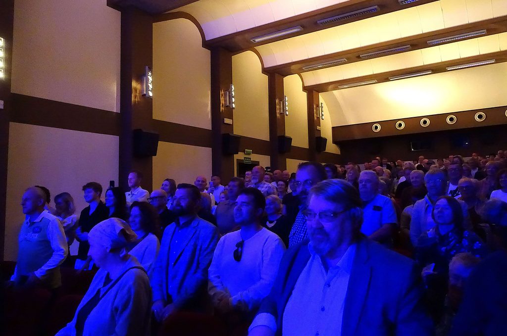 Ludzie stojący w widowni kina lub teatru, oświetleni niebieskim światłem, jasne ściany z ciemnymi ramami i punkty świetlne na suficie.
