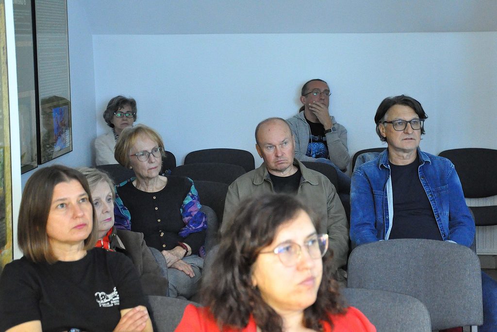 Grupa osób siedzących na szarych krzesłach w jasnym pomieszczeniu z białą ścianą, większość w okularach, ubrani w różne kolory, m.in. niebieską kurtkę i czarną bluzkę.