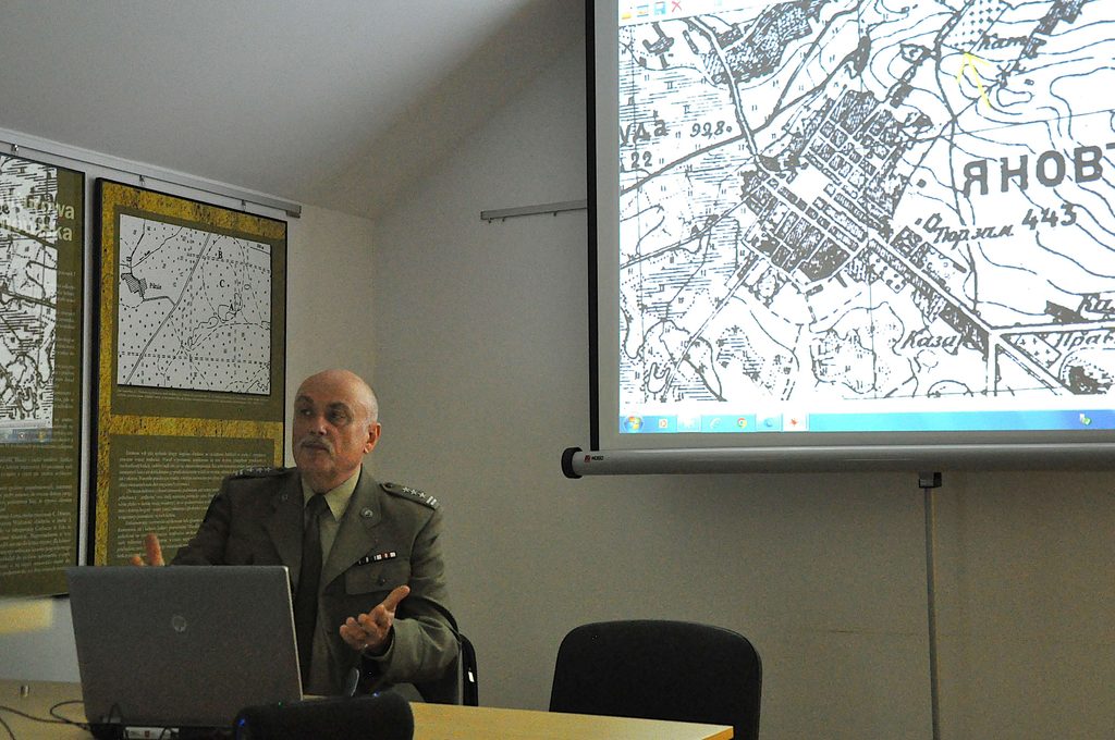 Mężczyzna w mundurze wojskowym siedzi przy stole z laptopem, za nim plansze z mapami, obok ekran projekcyjny z czarno-białą mapą.
