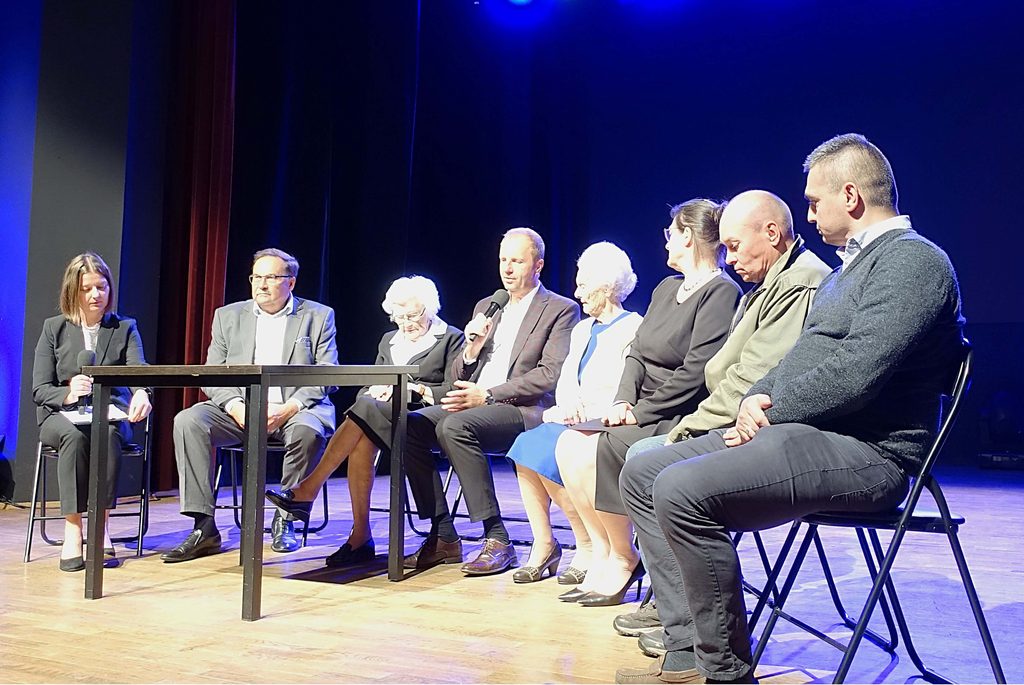 Osiem osób siedzących na krzesłach na scenie, jedna trzyma mikrofon, odzież formalna, ciemne tło z niebieskim oświetleniem.