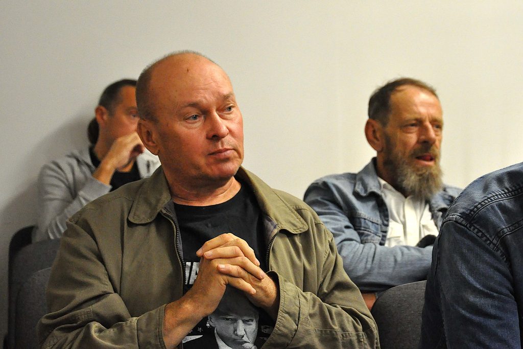Trzech mężczyzn na tle jasnej ściany, w centrum mężczyzna z łysiną i zieloną kurtką, po prawej brodaty mężczyzna w jasnoniebieskiej kurtce dżinsowej.
