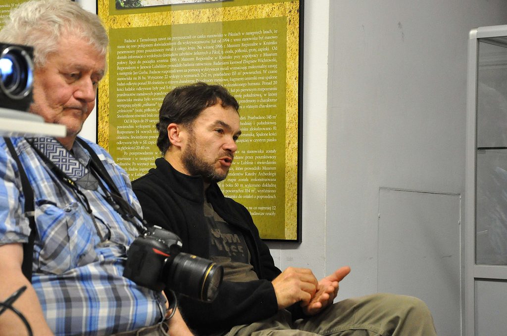 Dwóch mężczyzn siedzi na krzesłach, jeden w koszuli w kratę z aparatem fotograficznym, drugi w czarnej bluzie, za nimi zielono-żółty plakat z tekstem.