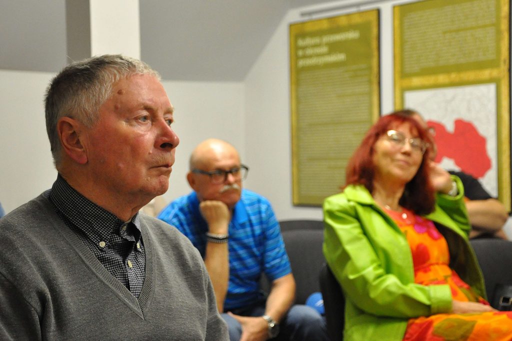 Trzy osoby siedzące w pokoju, mężczyzna z siwymi włosami w szarej bluzie, starszy mężczyzna w niebieskiej koszulce w paski, kobieta z rudymi włosami w zielonej marynarce i kolorowej sukience.