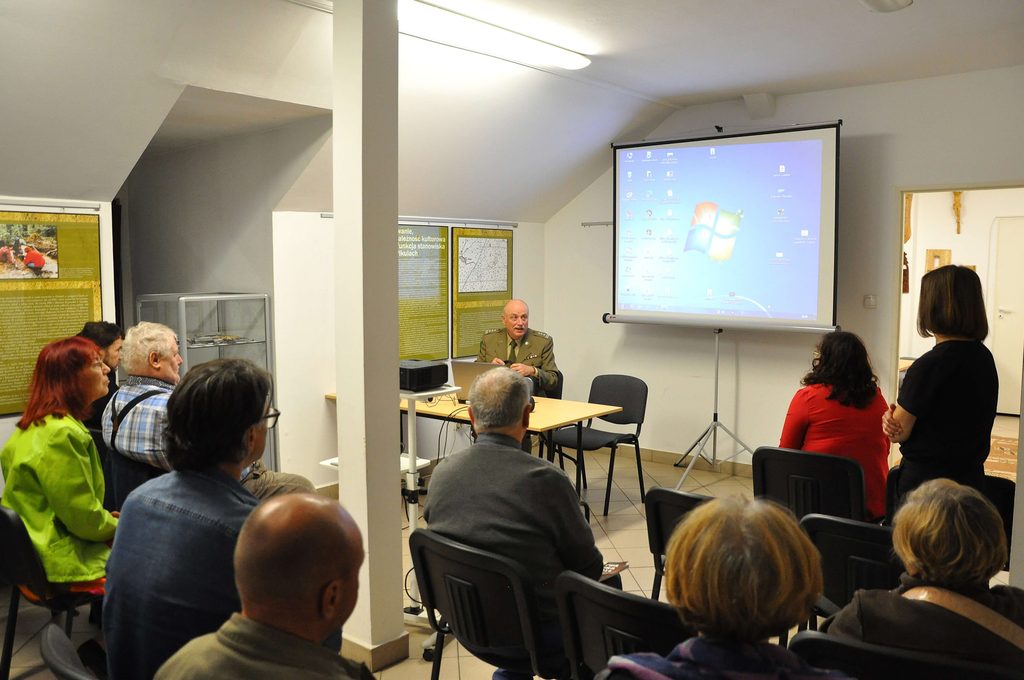 Sala konferencyjna z projekcją ekranu, uczestnicy siedzący i stojący, mężczyzna w mundurze przy stole, biała ściana, tablice informacyjne, jasne oświetlenie.