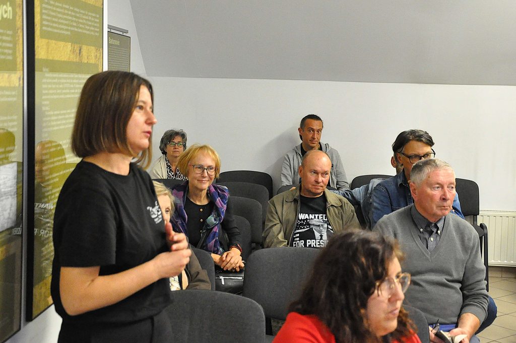 Grupa siedzących osób w pomieszczeniu z białymi ścianami, na pierwszym planie kobieta w czarnej koszulce, na ścianie po lewej tablice informacyjne.