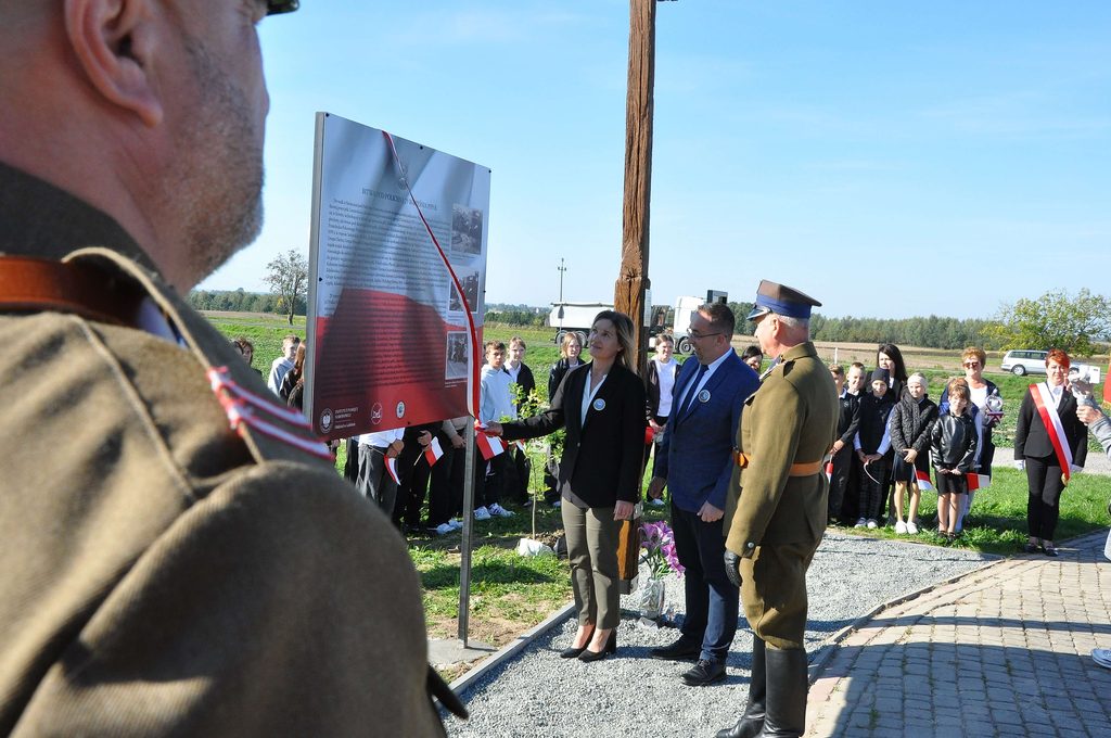 Grupa ludzi na zewnątrz, mężczyzna w mundurze wojskowym, kobieta i mężczyzna odsłaniają tablicę z tekstem i zdjęciami, w tle dzieci i dorośli, niebo bezchmurne.
