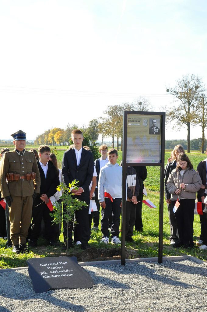 Grupa osób stojących na zielonym polu przy tablicy pamiątkowej i kamieniu z napisem, niebo jasne, drzewa z żółtymi liśćmi w tle.