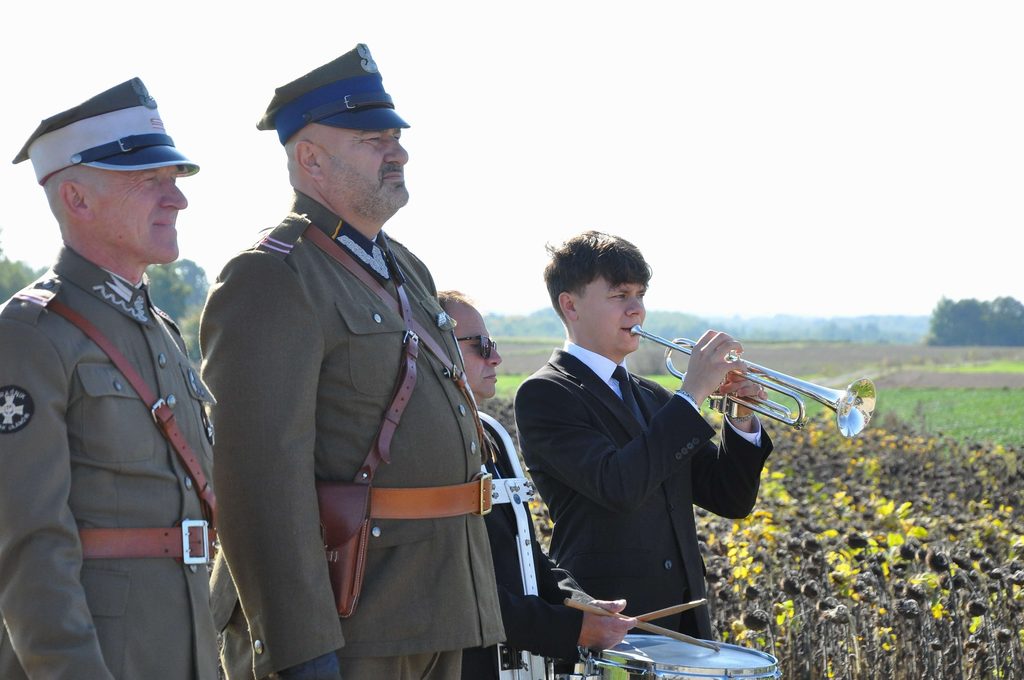 Trzech mężczyzn w mundurach wojskowych i młody mężczyzna w czarnym garniturze grający na trąbce na tle pola słoneczników.