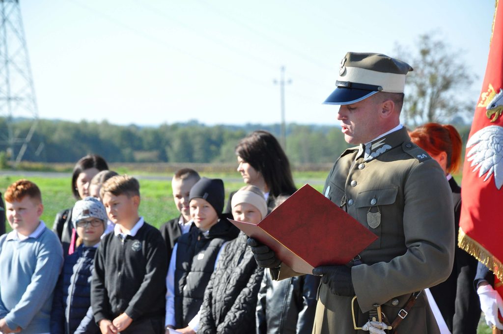 Mężczyzna w mundurze wojskowym i czapce czyta z czerwonego folderu, obok stoi grupa dzieci i młodzieży, w tle czerwona flaga z orłem i zielone pole.