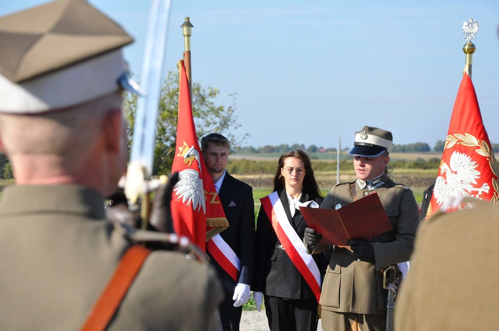 Trzech ludzi w mundurach i biało-czerwonych szarfach, czerwone flagi z godłem Polski, tło polna droga, drzewa i pole, niebieskie niebo.