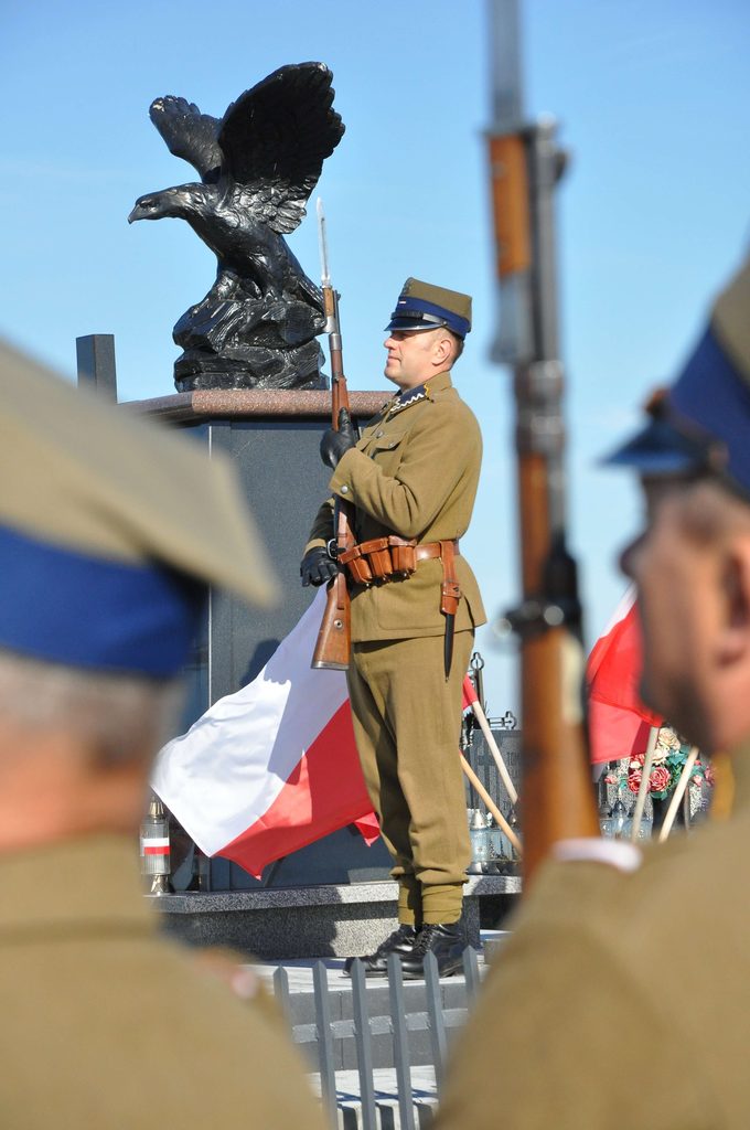 Żołnierz w historycznym mundurze z bronią na tle pomnika orła i flagi Polski, fragmenty innych żołnierzy w mundurach na pierwszym planie.