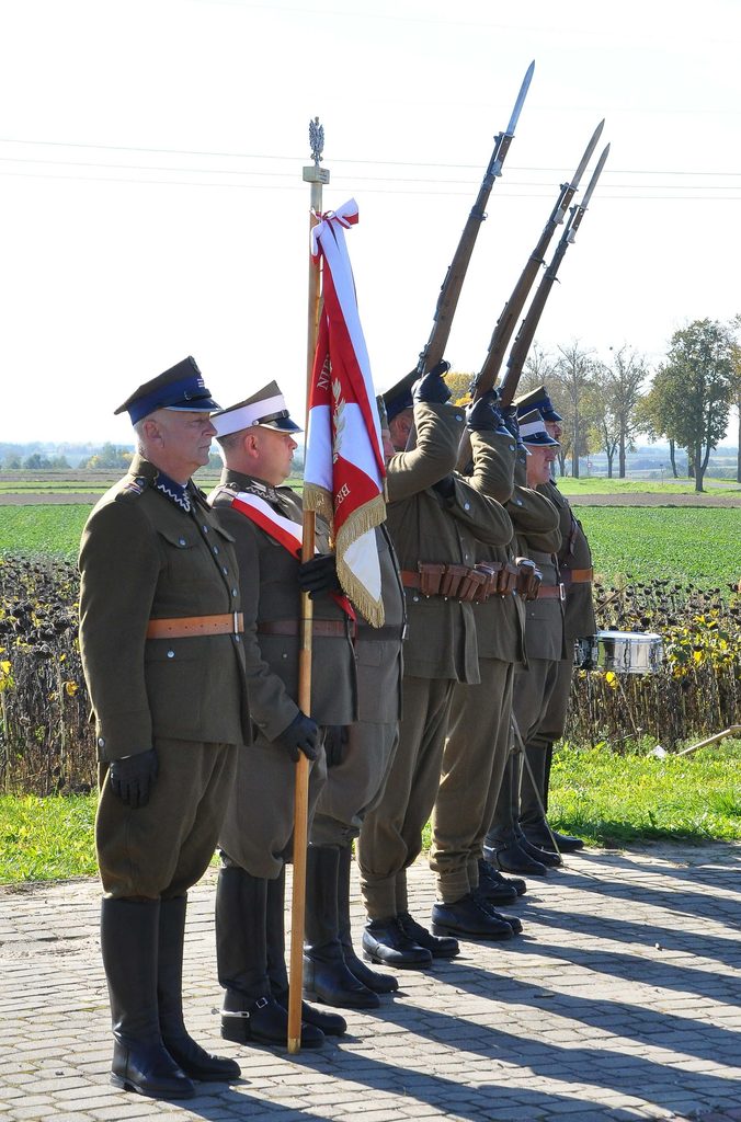 Mężczyźni w mundurach wojskowych w zielonym kolorze, trzymający sztandar i karabiny z bagnetami, stojący na chodniku obok pola i drzew.