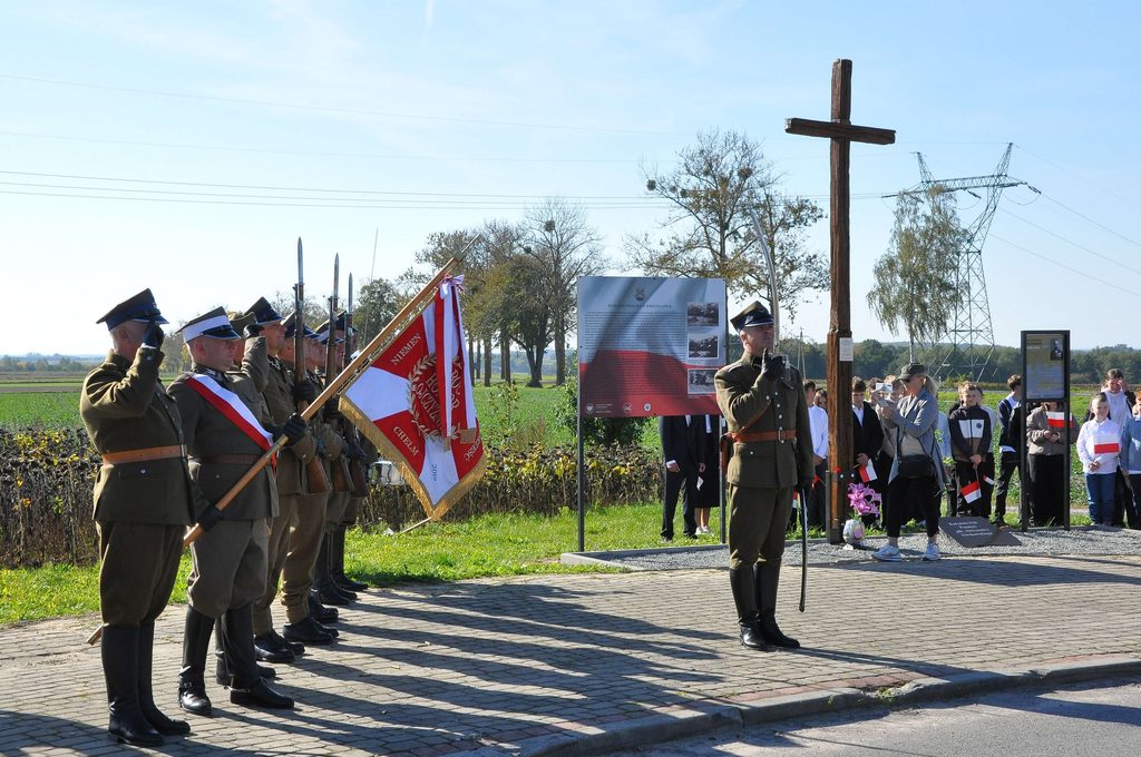 Żołnierze w mundurach z szablami i flagą, drewniany krzyż, tablice informacyjne, grupa ludzi z małymi flagami, drzewa i pola w tle pod niebieskim niebem.