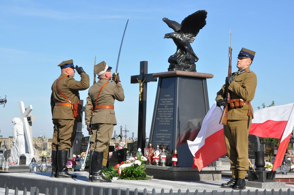 Trzech żołnierzy w mundurach historycznych przy pomniku z orłem, krzyżem i kwiatami, na tle niebieskiego nieba, z polskimi flagami.