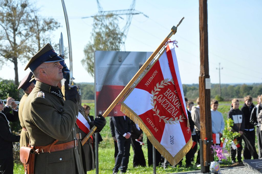 Mężczyzna w wojskowym mundurze z granatowo-złotym czapką trzymający sztandar biało-czerwony z złotymi frędzlami, w tle ludzie i tablica informacyjna.