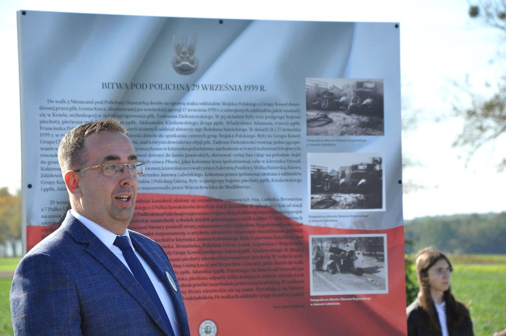 Mężczyzna w okularach i granatowym garniturze stoi przed tablicą informacyjną o bitwie pod Policzną z 1939 roku, w tle kobieta i drzewa na niebieskim niebie.