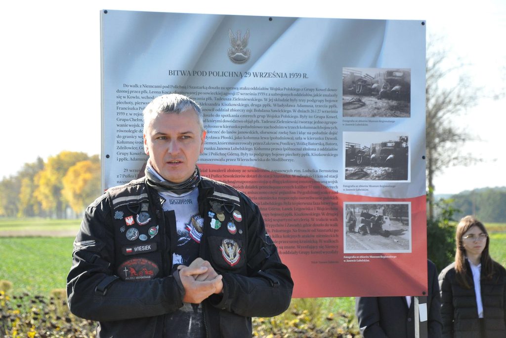 Mężczyzna w czarnej kurtce z naszywkami stoi na tle tablicy informacyjnej o bitwie pod Polichną, w tle zieleń i rozmyte postacie oraz drzewa.