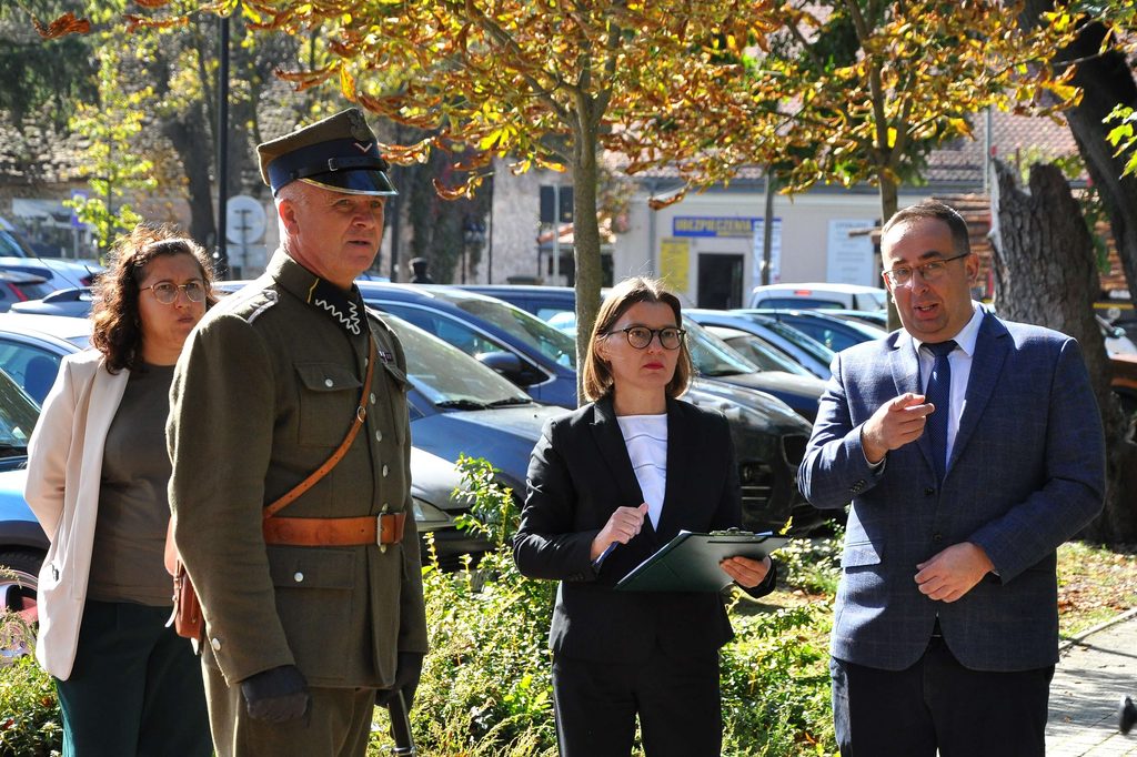 Cztery osoby na tle jesiennych drzew i zaparkowanych samochodów, mężczyzna w mundurze wojskowym, dwie kobiety w okularach i mężczyzna w garniturze.