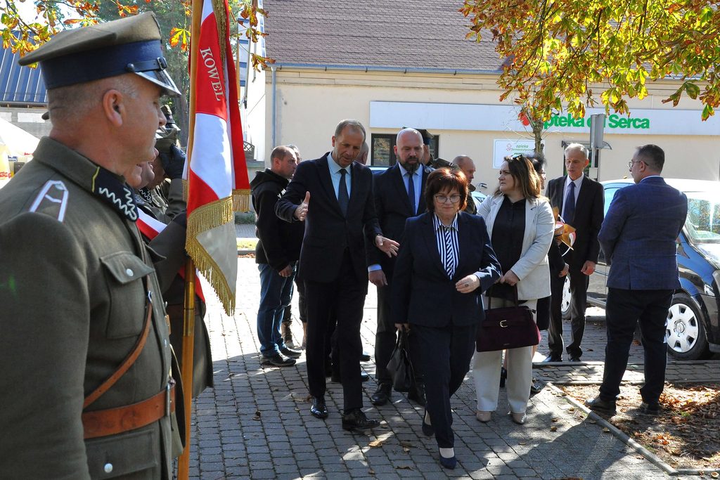Grupa ludzi na chodniku, mężczyzna w mundurze wojskowym trzyma flagę z napisem, jesienne liście na drzewach, budynek z napisem "Apteka od Serca" w tle.