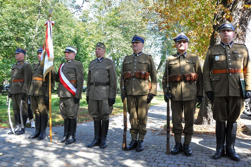 Siedmiu mężczyzn w wojskowych mundurach z bronią, stojących na brukowanym chodniku, w tle drzewa o zielonych i żółtych liściach. Jeden trzyma sztandar z białą i czerwoną wstęgą.