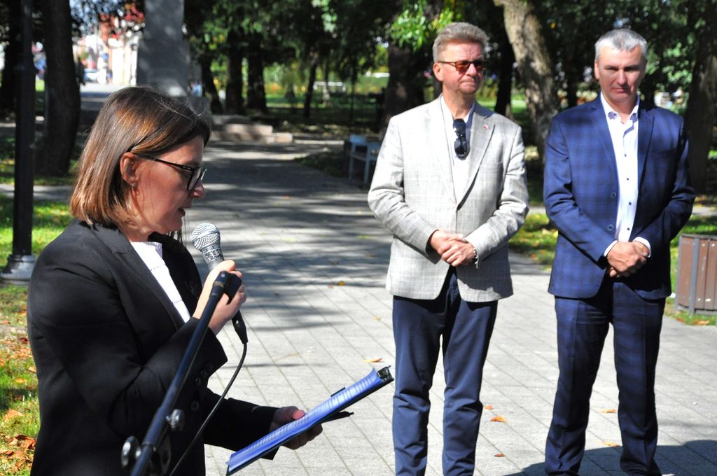 Kobieta z krótkimi brązowymi włosami i okularami trzyma mikrofon i kartkę, obok dwóch mężczyzn w garniturach stojących na brukowanym chodniku w parku.