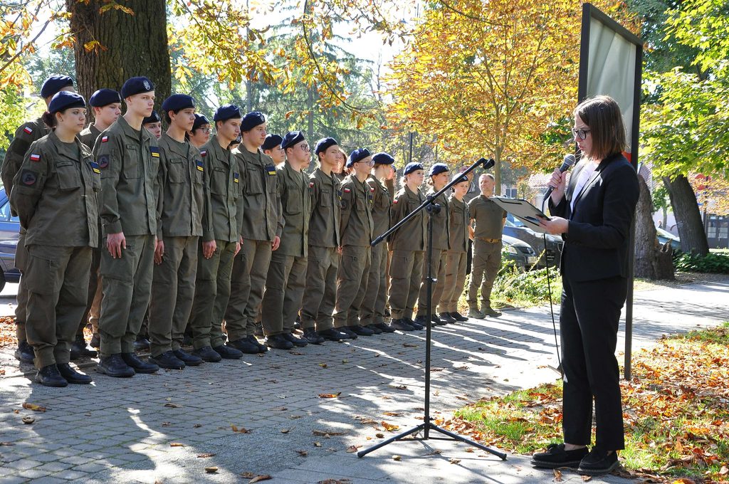 Grupa młodych osób w mundurach wojskowych i beretach stoi na tle jesiennych drzew, kobieta w czarnym garniturze czyta z kartki przy mikrofonie na stojaku.